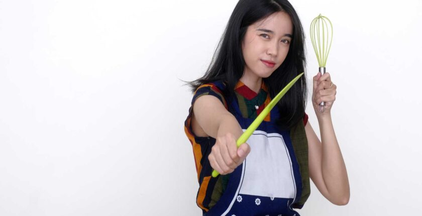 Heute lernen, morgen handeln - Ausbildung und Karriere - A young woman wearing an apron holds a spatula and a whisk in a serious expression